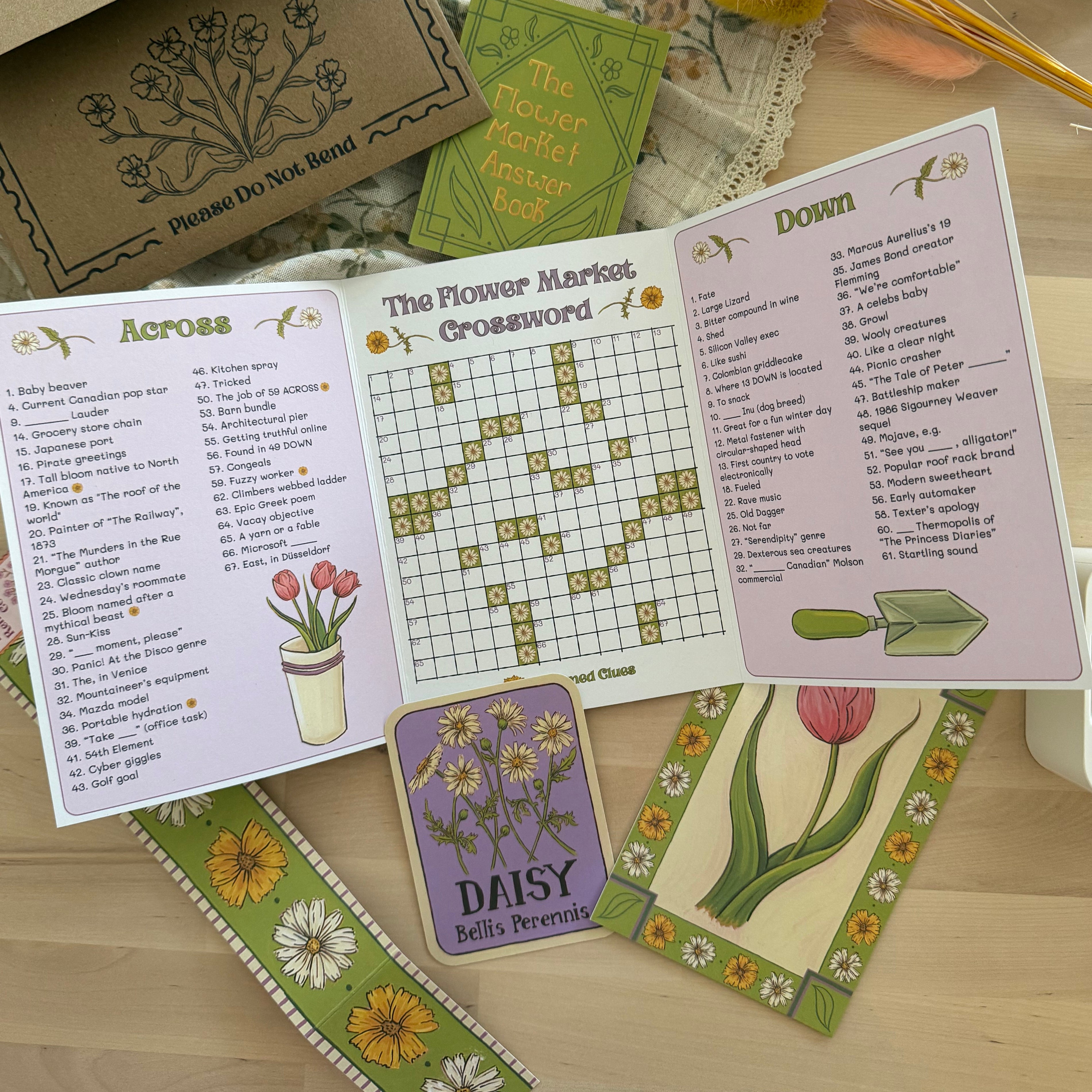 A floral-themed snail mail club crossword puzzle book set displayed on a wooden tabletop with decorative dried flowers and fabric. The set includes an open spread showing Across and Down clues, a central 'The Flower Market' crossword page, a purple 'Daisy Bell's Preferences' card, and several illustrated cards with tulips and flowers.