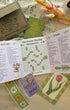 A floral-themed crossword puzzle game set displayed on a light wooden table. The set includes an open booklet showing 'Across' and 'Down' clues with a flower crossword grid, a green 'The Flower Market' booklet, a brown envelope with printed floral designs, and several decorative cards featuring tulips and daisies.
