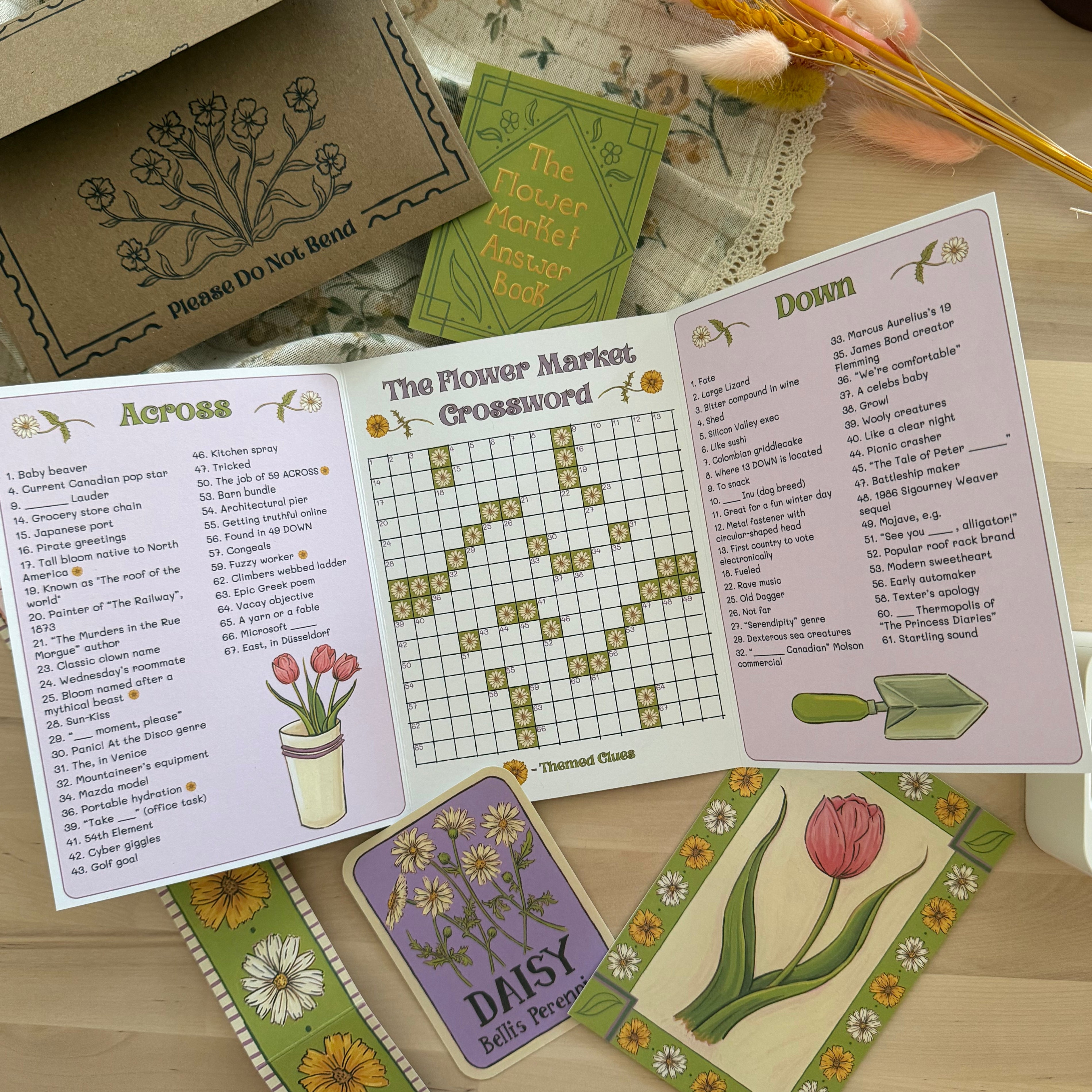 A floral-themed crossword puzzle game set displayed on a light wooden table. The set includes an open booklet showing 'Across' and 'Down' clues with a flower crossword grid, a green 'The Flower Market' booklet, a brown envelope with printed floral designs, and several decorative cards featuring tulips and daisies.