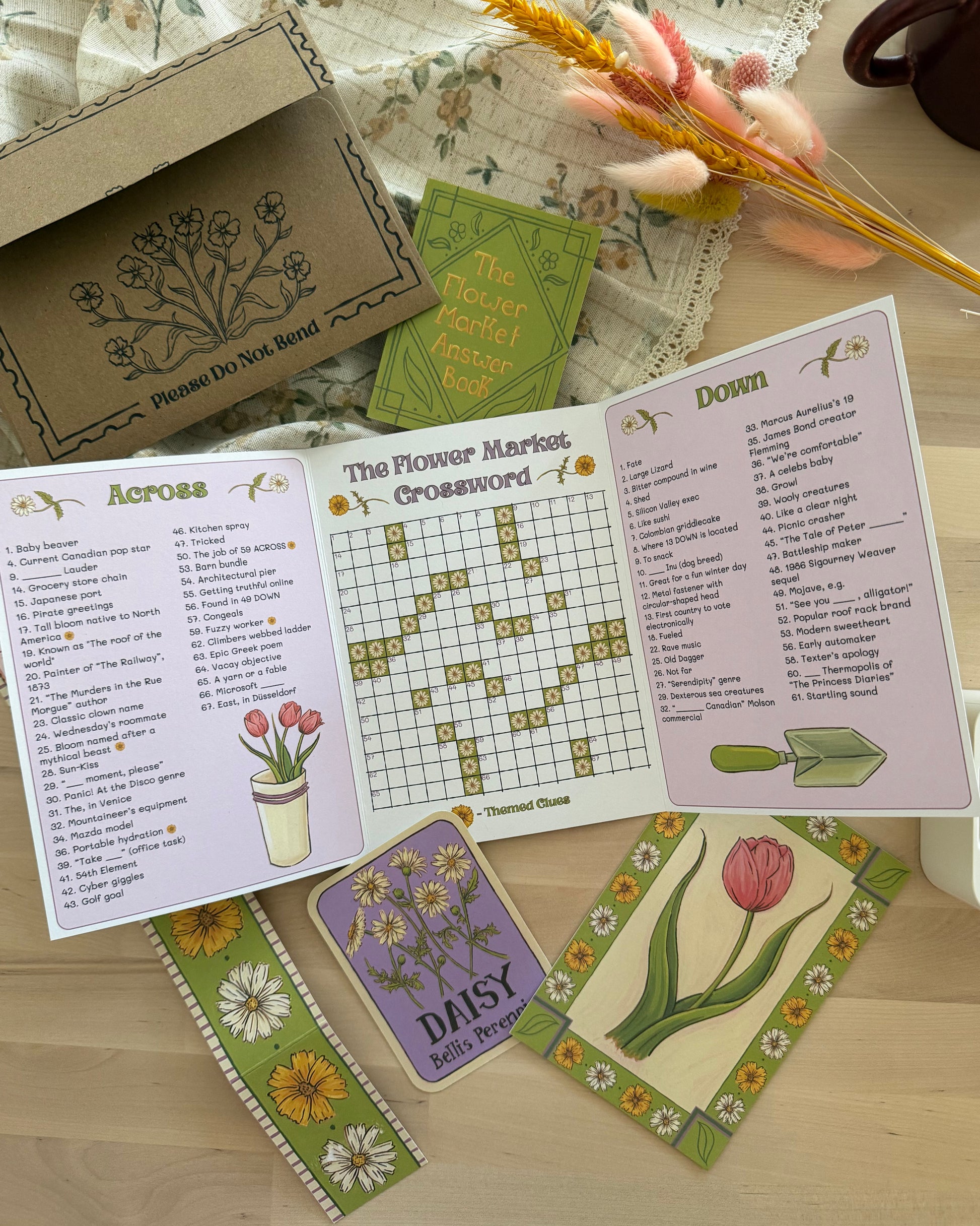 A floral-themed crossword puzzle game set displayed on a light wooden table. The set includes an open booklet showing 'Across' and 'Down' clues with a flower crossword grid, a green 'The Flower Market' booklet, a brown envelope with printed floral designs, and several decorative cards featuring tulips and daisies.