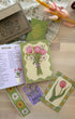 A floral-themed card set and recipe booklet displayed on a light wood tabletop. The set includes a brown kraft envelope with floral printing, a green 'The Flower Market Recipe Book', an open recipe page titled 'Across', a card with pink tulips, a purple card reading 'Daisy', and a green-bordered card with a single pink tulip.