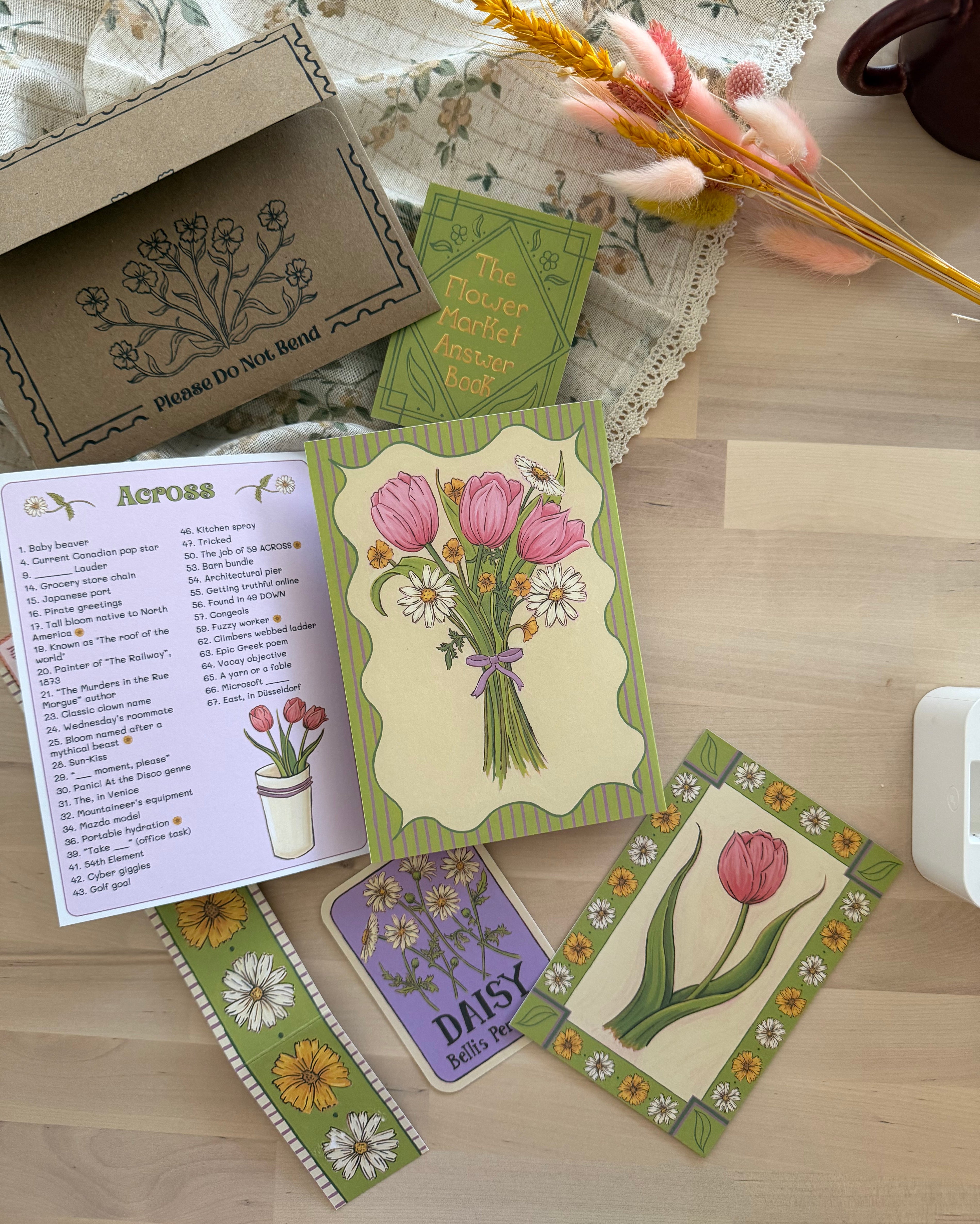 A floral-themed card set and recipe booklet displayed on a light wood tabletop. The set includes a brown kraft envelope with floral printing, a green 'The Flower Market Recipe Book', an open recipe page titled 'Across', a card with pink tulips, a purple card reading 'Daisy', and a green-bordered card with a single pink tulip.
