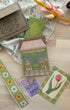 A styled flat-lay of a floral-themed stationery set on a light wood surface, featuring a brown kraft paper box with botanical line art, a green patterned card, a colorful storefront illustration card, a purple card reading 'Daisy', a green floral belly band, and a card with a pink tulip. Decorative dried flowers, lace fabric, and a dark mug are arranged around the items.