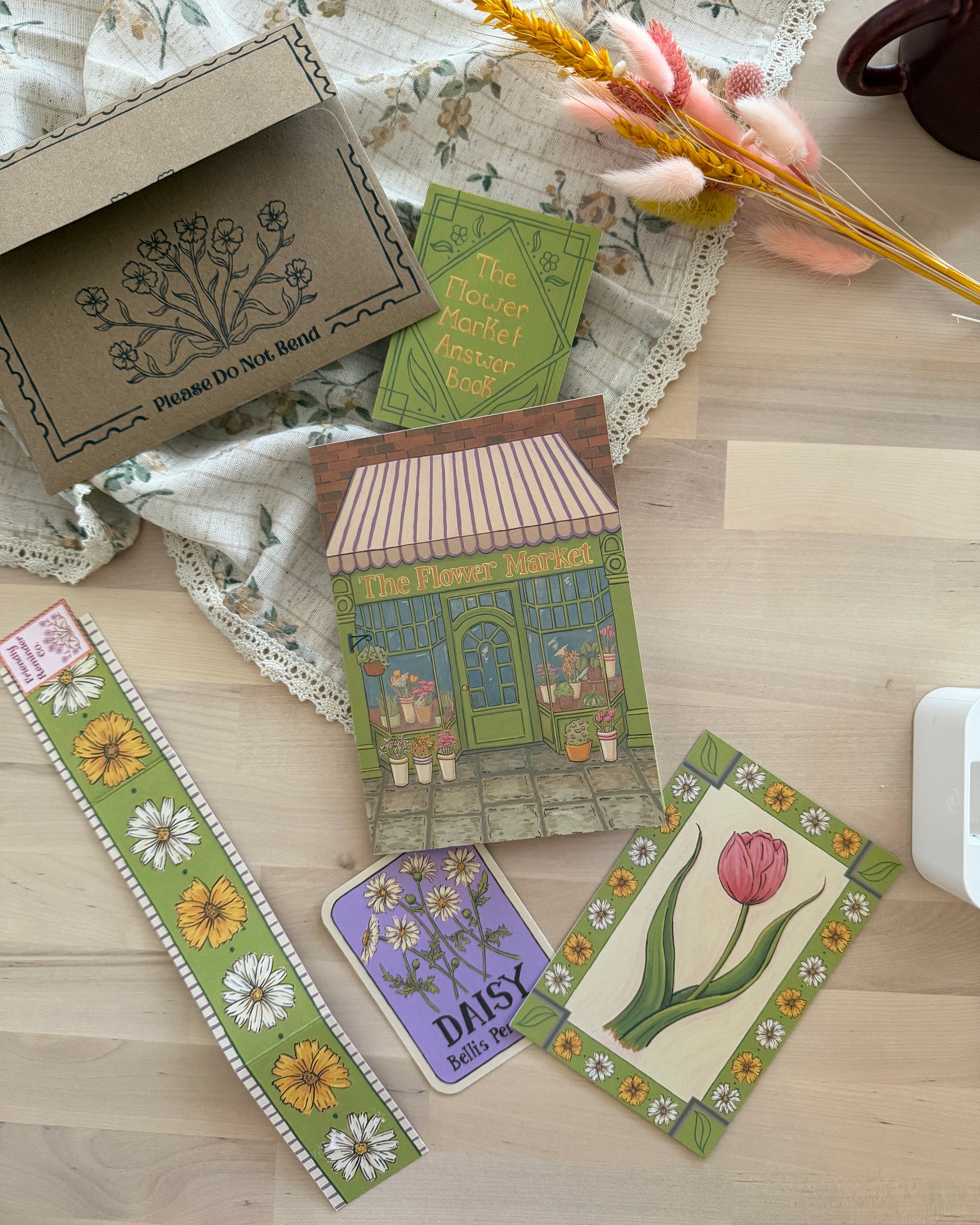 A styled flat-lay of a floral-themed stationery set on a light wood surface, featuring a brown kraft paper box with botanical line art, a green patterned card, a colorful storefront illustration card, a purple card reading 'Daisy', a green floral belly band, and a card with a pink tulip. Decorative dried flowers, lace fabric, and a dark mug are arranged around the items.