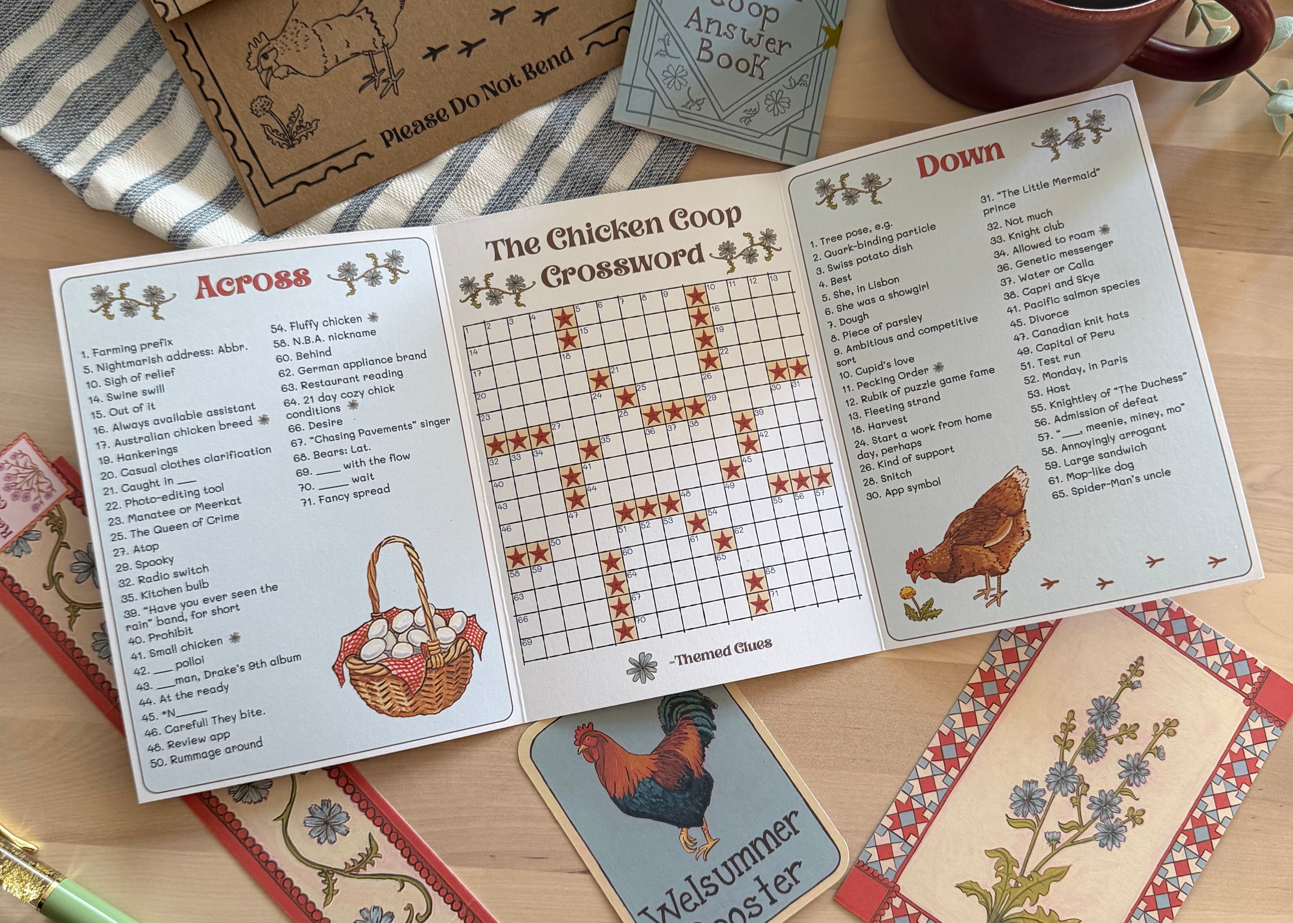 A rustic flat-lay of a chicken-themed word game set on a wooden table, featuring an open game booklet with clues, a card deck, a small envelope, a striped cloth, a mug of coffee, and decorative floral postcard.